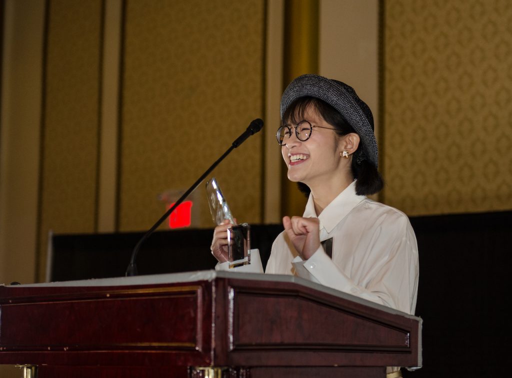 Student stands at podium while smiling and hold an award she has won