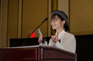 Student stands at podium while smiling and hold an award she has won