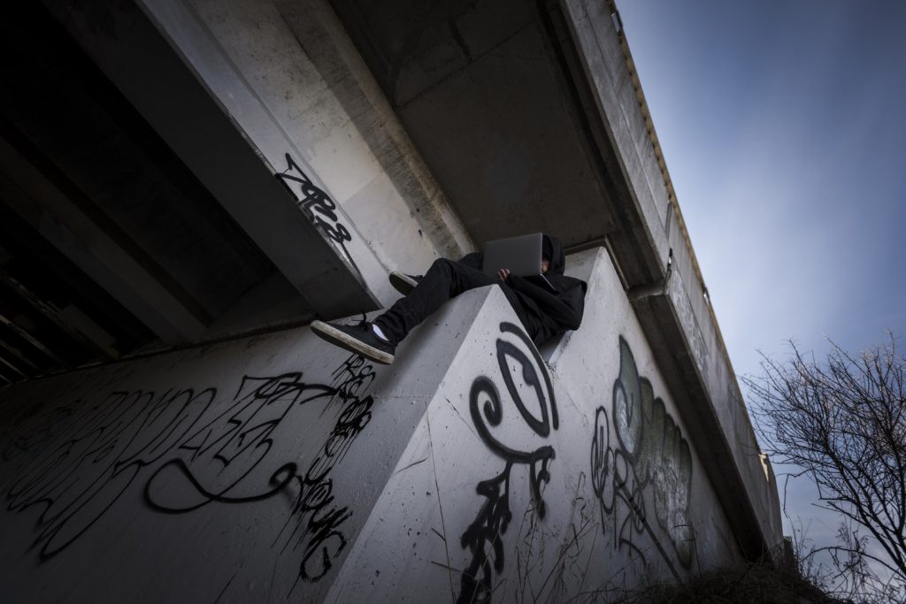 Man in black clothing sits on concrete highway under pass ledge holding laptop while his face is partially covered.