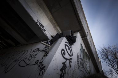 Man in black clothing sits on concrete highway under pass ledge holding laptop while his face is partially covered.