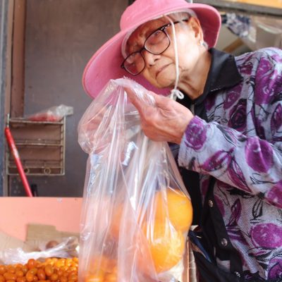 woman wearing hat at grocery store, picking out oranges and putting them into a plastic bag