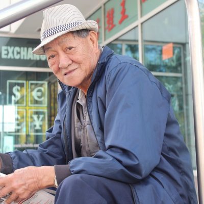 old man wearing a hat sits on the steps of Chinatown reading a newspaper