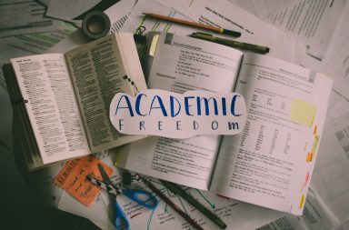 Photo of messy papers scattered throughout the frame. Writing utensils, tape and scissors are surrounding two open books. The words "Academic Freedom" are written on a piece of paper in blue marker with pencil traces around it.