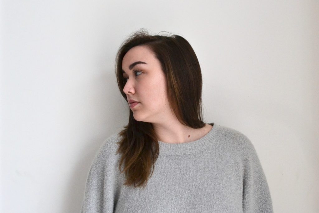 Portrait of female student in grey sweatshirt against wall with head turned left and looking away.