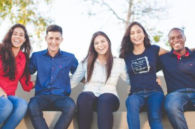 five college students sitting on a bench with their arms around eachother. they're smiling.